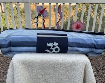 May include: A blue and white yoga mat bag with a navy blue pocket featuring a white Om symbol. The bag is lying on a white fluffy surface.