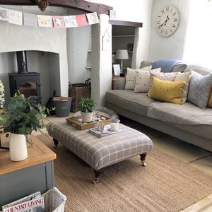 May include: A living room with a grey sofa, a grey and white plaid footstool, a jute rug, and a wood-burning stove. The room is decorated with throw pillows, a clock, and a garland of cards.