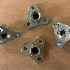 May include: Four silver metal threaded inserts with three prongs each. The inserts are designed to be used in wood or other materials to create a threaded hole for attaching screws or bolts.