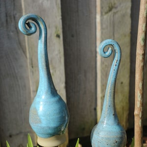 Può includere: Tre picchetti da giardino in ceramica turchese. Ogni picchetto presenta un corpo bulboso e una sommità curva e a spirale. Sono inseriti in piante verdi, con una staccionata di legno sullo sfondo. I picchetti sono alti circa 30 cm.