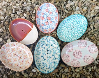 Könnte beinhalten: Fünf ovale Seifenschalen aus Keramik mit Abflusslöchern. Eine ist rot-weiß, eine hat ein rosa Blumenmuster, eine zeigt einen türkisfarbenen geometrischen Druck, eine hat ein blaues Blattdesign und eine ist mit einem roten Punktmuster versehen.