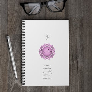 May include: A white spiral bound journal with a pink and white mandala design and the words "I Am" in a cursive font. The words "infinite, limitless, peaceful, spiritual, conscious" are printed below the mandala. The journal has a black and white Om symbol at the top.