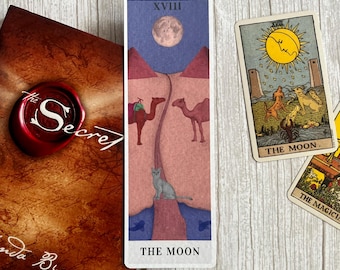 Könnte beinhalten: Eine Sammlung von Tarotkarten und ein Buch mit dem Titel "The Secret" sind auf einer hellen Holzoberfläche angeordnet. Die zentrale Karte, "The Moon", zeigt einen Vollmond, Kamele und eine Katze. Weitere Karten sind "The Sun" und "The Magician".