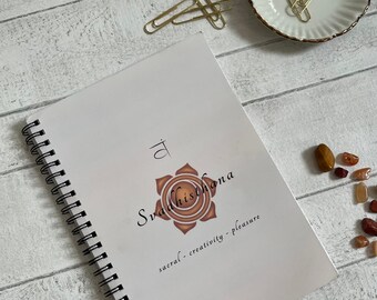 Könnte beinhalten: Ein weißes Spiralnotizbuch mit dem Wort "Svadhisthana" und einem orangefarbenen Chakra-Symbol auf dem Cover. Der Text "sacral - creativity - pleasure" steht darunter. Goldene Büroklammern und kleine Steine sind ebenfalls zu sehen.