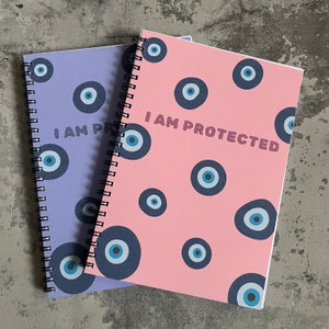 Op de afbeelding: Twee spiraalgebonden notitieboekjes met een blauw en wit boze oogpatroon. Het roze notitieboekje zegt "I AM PROTECTED" in vetgedrukte letters.