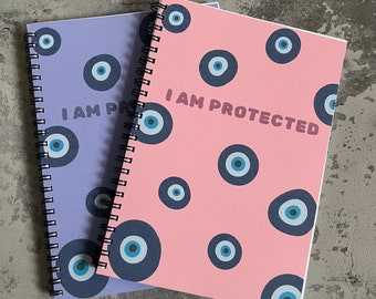 Könnte beinhalten: Zwei spiralgebundene Notizbücher mit einem blauen und weißen bösen Augenmuster. Das rosa Notizbuch sagt "I AM PROTECTED" in fetter Schrift.