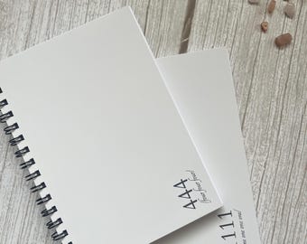 Könnte beinhalten: Zwei weiße Notizbücher mit schwarzer Spiralbindung. Das obere Notizbuch zeigt den schwarzen Text 444 /four four four/. Das untere Notizbuch zeigt den schwarzen Text 1111 /one one one one/. Beide Notizbücher haben schlichte weiße Einbände.