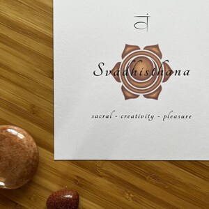 May include: A white card with a brown lotus flower illustration and the Sanskrit word "Svadhisthana" written in black. The text "sacral - creativity - pleasure" is written below the flower.