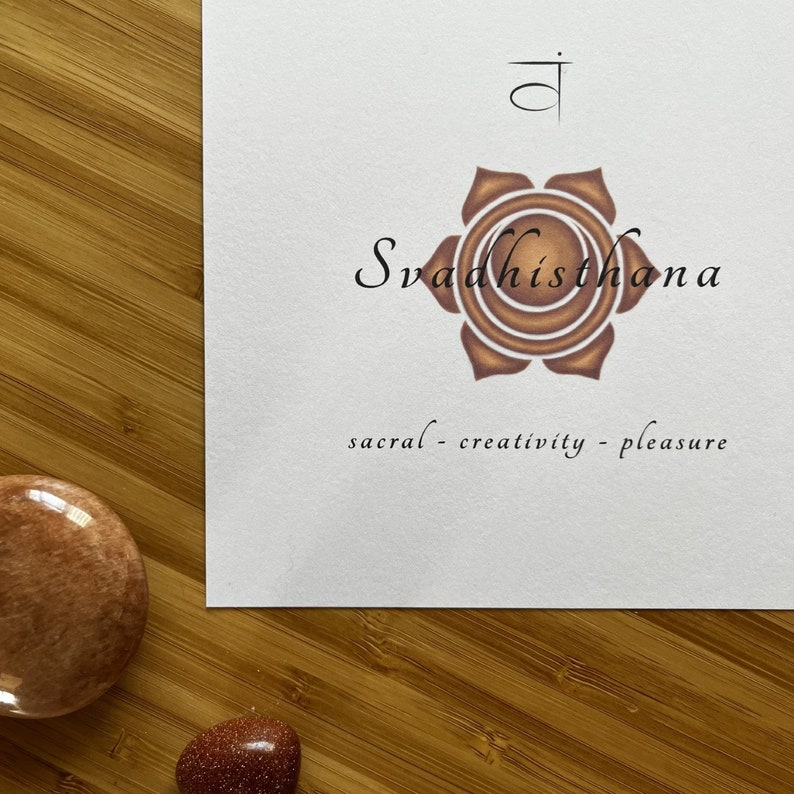 May include: A white card with a brown lotus flower illustration and the Sanskrit word "Svadhisthana" written in black. The text "sacral - creativity - pleasure" is written below the flower.