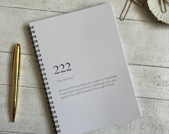 Könnte beinhalten: Ein weißes Spiralnotizbuch mit der Zahl 222 und dem Text "two two two" auf dem Cover. Der Text unter der Zahl lautet "think of things that you want to manifest, trust that everything is working out, you are in the right place at the right time."