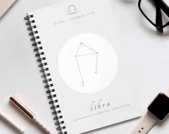 Könnte beinhalten: Ein weißes Spiralnotizbuch mit einem Waage-Sternzeichen-Design. Das Cover zeigt das Waage-Symbol, die Konstellation und das Wort "Waage" mit den Eigenschaften "Diplomatisch, Sozial, Anmutig". Ein Stift, eine Uhr und eine Brille sind ebenfalls zu sehen.