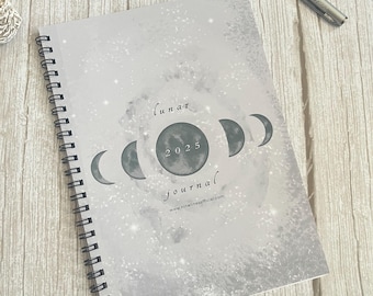 Könnte beinhalten: Ein spiralgebundenes Notizbuch mit hellgrauem Einband und Mondthema. Der Einband zeigt ein Mondphasen-Design mit dem Text "lunar journal 2025" und der Website-Adresse. Ein silberner Stift und eine Geode sind ebenfalls zu sehen.