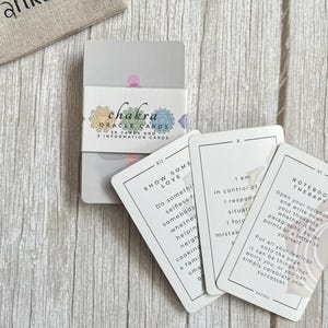 Könnte beinhalten: Ein Chakra-Orakelkarten-Deck mit einer weißen Papierbanderole, die bunte Chakrasymbole und den Text Chakra Oracle Cards 38 cards and 3 information cards zeigt. Drei einzelne Karten sind aufgefächert und zeigen Text sowie römische Ziffern.