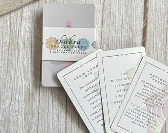Könnte beinhalten: Ein Chakra-Orakelkarten-Deck mit einer weißen Papierbanderole, die bunte Chakrasymbole und den Text Chakra Oracle Cards 38 cards and 3 information cards zeigt. Drei einzelne Karten sind aufgefächert und zeigen Text sowie römische Ziffern.