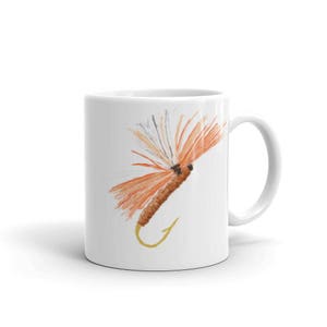 May include: A white ceramic mug with a colorful fishing fly illustration. The fly has orange, brown, and white feathers. The mug has a handle on the right side.