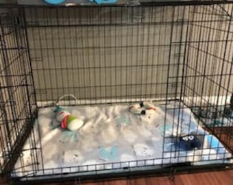 May include: A black metal dog crate with two open doors. Inside, a patterned blanket with blue and white designs covers the floor. Toys, including a plush toy and a chew toy, are inside the crate.