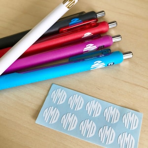 Puede incluir: Una hoja de pegatinas de monograma blancas con las letras "VMD" en un círculo. Hay 10 pegatinas en la hoja. También hay cinco bolígrafos con las mismas pegatinas de monograma. Los bolígrafos son negros, rojos, rosas, azules y blancos.