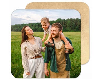 May include: A square coaster featuring a photograph of a family in a green field. A person in a beige shirt and white trousers stands next to another person in a green shirt and tan overalls carrying a small child on their shoulders.