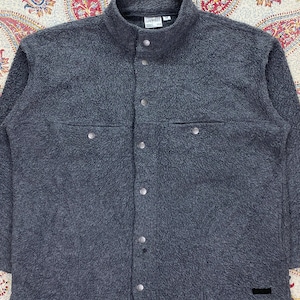 May include: A dark gray fleece jacket with a high collar and button-up front. The jacket has two chest pockets with button closures and a small black label near the bottom. The fabric has a textured, fuzzy appearance.