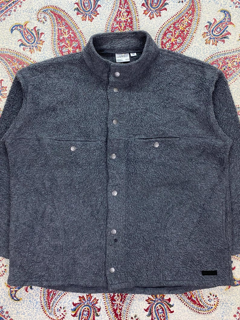 May include: A dark gray fleece jacket with a high collar and button-up front. The jacket has two chest pockets with button closures and a small black label near the bottom. The fabric has a textured, fuzzy appearance.