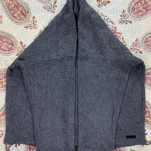 May include: A dark gray, hooded scarf made of a soft, textured material. The scarf is folded in a triangular shape, with a hood at the top and a rectangular label near the bottom. The scarf is displayed on a patterned fabric.