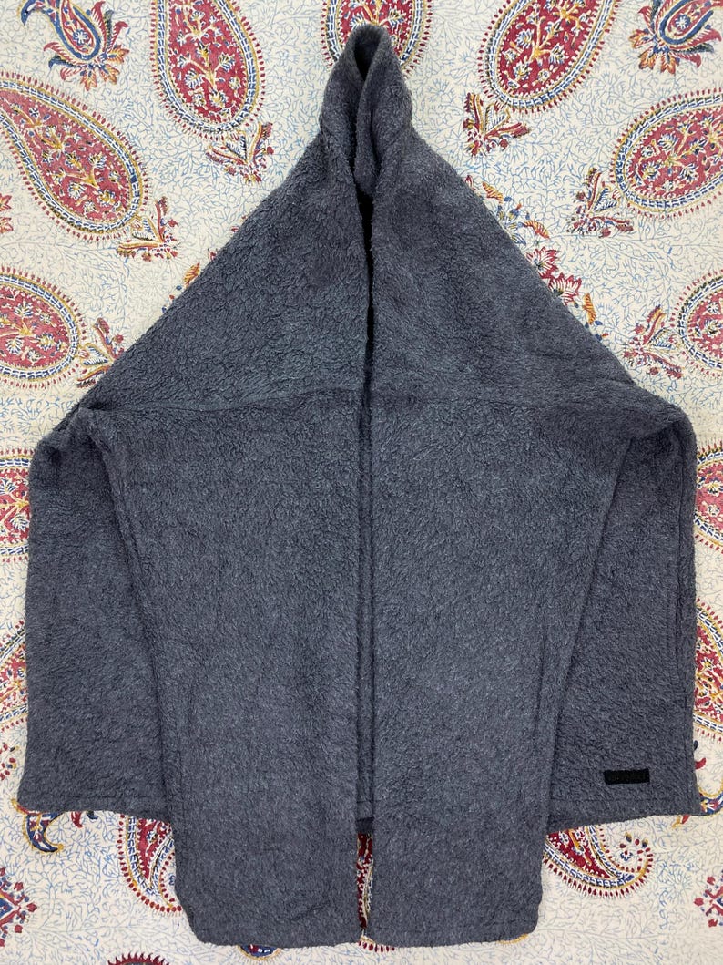 May include: A dark gray, hooded scarf made of a soft, textured material. The scarf is folded in a triangular shape, with a hood at the top and a rectangular label near the bottom. The scarf is displayed on a patterned fabric.