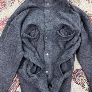 May include: A gray fleece jacket with a high collar and snap closures. The jacket has two large front pockets and a soft, textured appearance. The sleeves are long and the overall design is casual and warm.