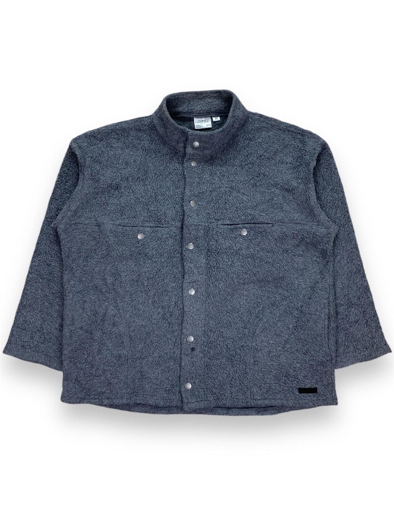 May include: A dark gray fleece jacket with a button-up front, two chest pockets, and a stand-up collar. The jacket has a textured appearance and a small black label near the bottom.