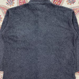 May include: A charcoal gray fleece jacket with a high collar. The jacket has a soft, textured appearance. The background features a patterned fabric with red, blue, and yellow paisley designs.