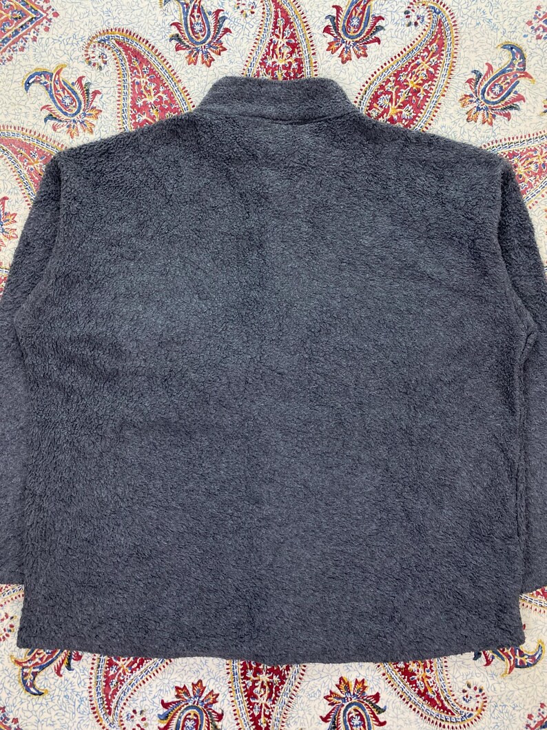 May include: A charcoal gray fleece jacket with a high collar. The jacket has a soft, textured appearance. The background features a patterned fabric with red, blue, and yellow paisley designs.