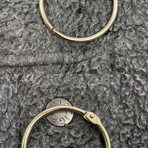 May include: Two silver-colored binder rings with a hinged opening, set against a textured gray background. Small, circular metal pieces with the letters "GR" and "M" are also visible.