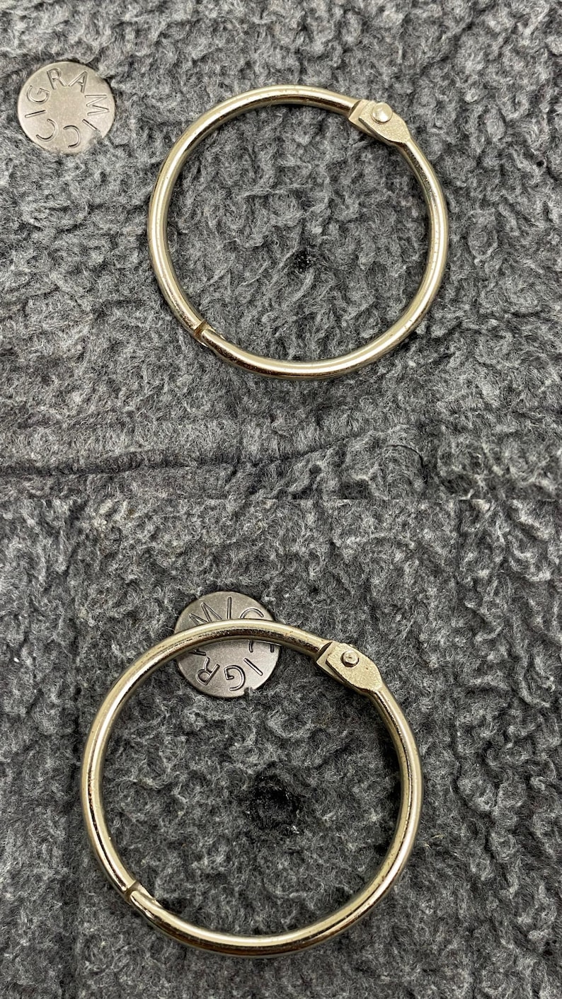 May include: Two silver-colored binder rings with a hinged opening, set against a textured gray background. Small, circular metal pieces with the letters "GR" and "M" are also visible.