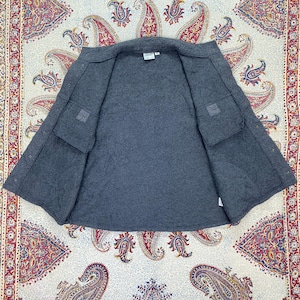 May include: A gray, open jacket with two front pockets and a quilted interior. The jacket is laid flat, showcasing its interior lining and construction. The garment appears to be made of a thick, warm material, suitable for cooler weather.