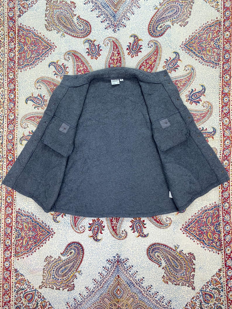 May include: A gray, open jacket with two front pockets and a quilted interior. The jacket is laid flat, showcasing its interior lining and construction. The garment appears to be made of a thick, warm material, suitable for cooler weather.