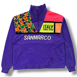 Puede incluir: Chaqueta de forro polar morada con cuello alto y cierre de media cremallera. La chaqueta presenta un diseño de bloques de color con un panel rosa neón y naranja, y una sección estampada con formas verdes, naranjas y amarillas. Se ve el texto "SANMARCO" y "ITALY SINCE 1948".