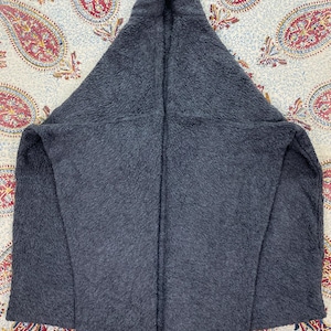 May include: A dark gray, hooded fleece jacket. The jacket is made of a soft, textured material and features a large hood and open front. The jacket is laid flat on a patterned surface.