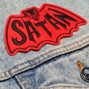 Könnte beinhalten: Roter gestickter Aufnäher in Form einer Fledermaus mit dem Wort "SATAN" in schwarzen Buchstaben. Die Fledermaus hat einen kleinen Teufelskopf in der Mitte.