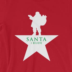 May include: Red t-shirt featuring a white silhouette of Santa Claus standing on a white star with the text "SANTA I BELIEVE" in green letters.