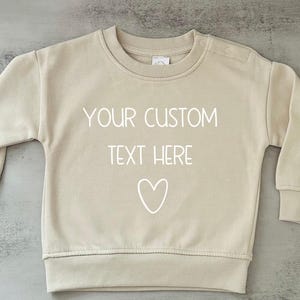 May include: A beige sweatshirt with a white heart and the text "YOUR CUSTOM TEXT HERE" printed on the front.