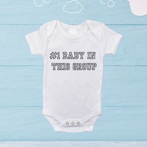 Peut inclure: Body de bébé blanc avec le texte "#1 BABY IN THIS GROUP" en lettres noires. Le body a des manches courtes et trois boutons-pression en bas. Le fond est bleu clair avec des formes de nuages blancs et le texte "Cloud Nine Co".