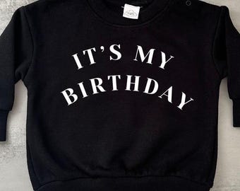 May include: Black crew neck sweatshirt with the text "IT'S MY BIRTHDAY" in white block letters. The sweatshirt has long sleeves and a ribbed hem and cuffs. There are two buttons on the left shoulder.