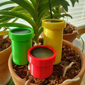 May include: Three 3D printed pipes in green, yellow, and red, resembling the iconic pipes from the Super Mario video game series. The pipes are sitting in a terracotta pot filled with brown mulch.