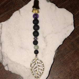 May include: A gold leaf charm with a black lava rock and purple gemstone beaded necklace. The necklace has a gold clip at the top.