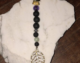 May include: A gold leaf charm with a black lava rock and purple gemstone beaded necklace. The necklace has a gold clip at the top.