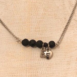 May include: A silver chain necklace with a charm that says "E" and a small heart. The charm is attached to a strand of black lava beads.