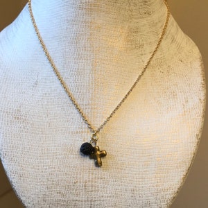 May include: A gold chain necklace with a small cross charm and a black lava rock bead.
