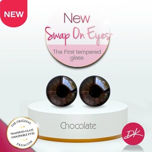 May include: Two brown glass eyes for doll making. The eyes are on a white platform with the words "New Swap On Eyes" and "The First tempered glass" in pink and gold lettering. The words "Chocolate" and "DK" are also visible.