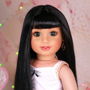 May include: A doll with long black hair and bangs, wearing a white tank top with a black bow, striped shorts, and white socks. The doll has green eyes and is wearing gold hoop earrings.