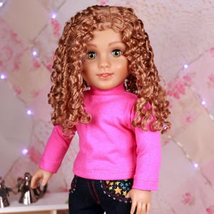May include: A doll with curly brown hair, wearing a pink long-sleeved shirt and dark blue jeans with star accents. The doll is also wearing pink shoes. The background has a floral pattern and a wooden floor.