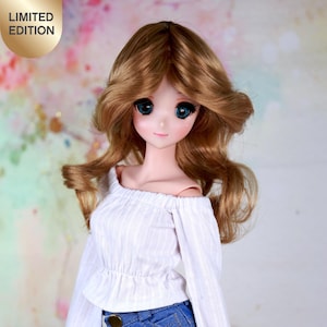 Könnte beinhalten: Eine Puppe trägt ein weißes, schulterfreies Oberteil mit langen Ärmeln und blaue Jeans-Shorts mit weißem Spitzenbesatz. Die Puppe hat langes blondes Haar und blaue Augen. Der Hintergrund ist ein verschwommenes rosa und grünes Aquarellmuster.